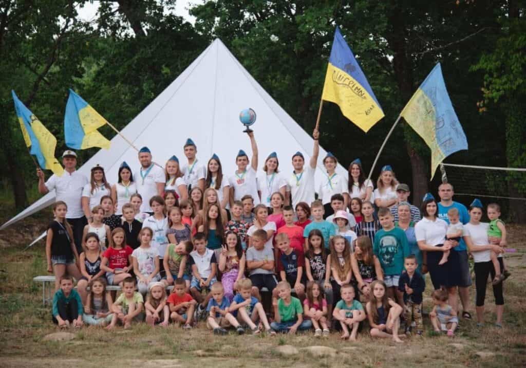 A large group of children and leaders with Ukrainian flags in front of a white tent