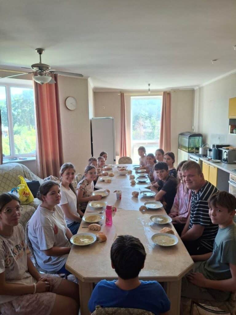 Children and teens sit together at a long table sharing a meal of soup and bread